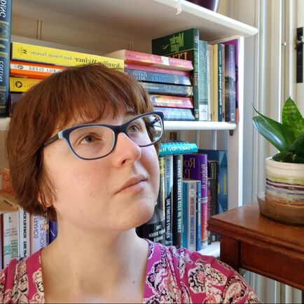 white woman with short brown hair, green eyes, and blue glasses in front of a bookshelf